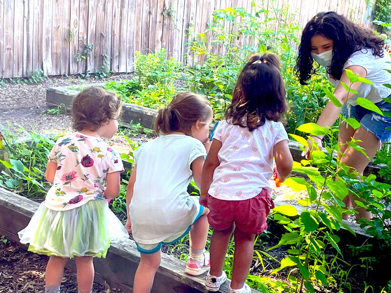 Children standing on a garden box in front of a woman wearing a face mask 
