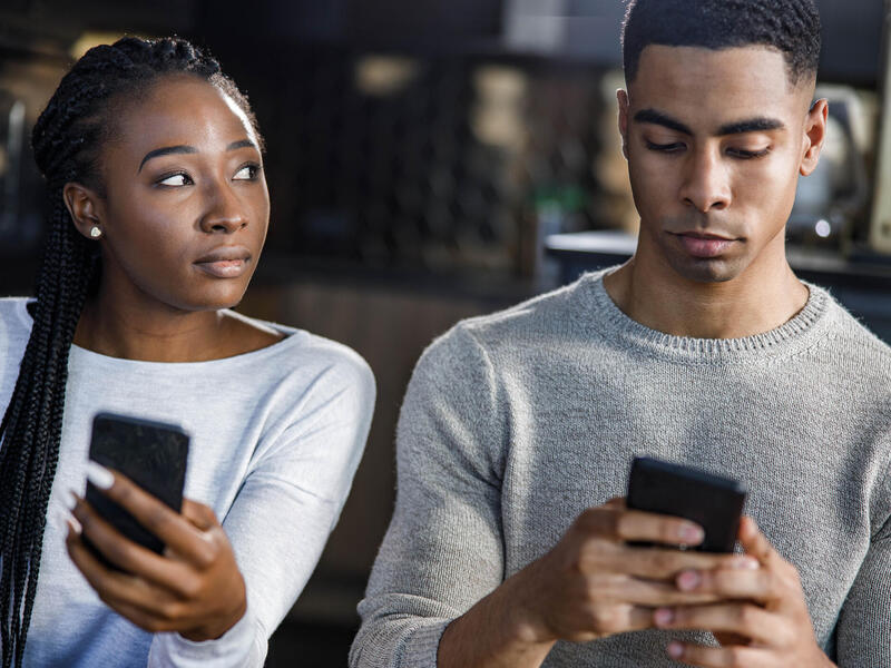 A photo of a man and woman sitting next to each other. Both people are holding cell phones, but the woman is looking at the man to her right supsicously while the man is looking at his phone. 