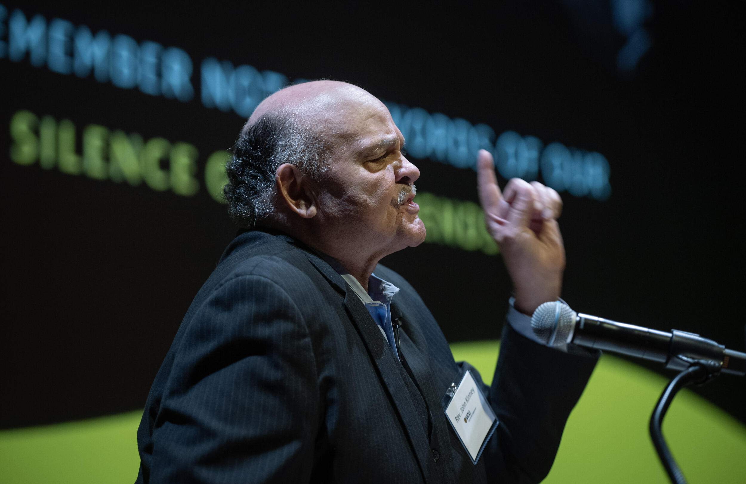 A photo of a man from the chest up speaking into a microphone. 