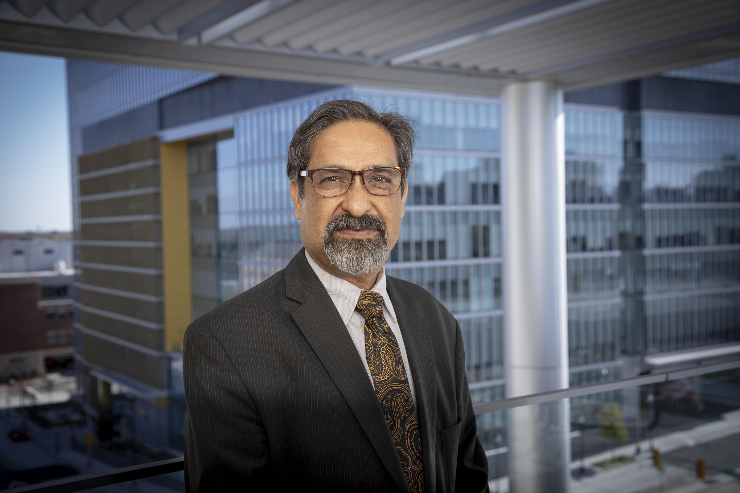 A photo of a man wearing a suit and tie from the chest up. He is standing in front of a window. 