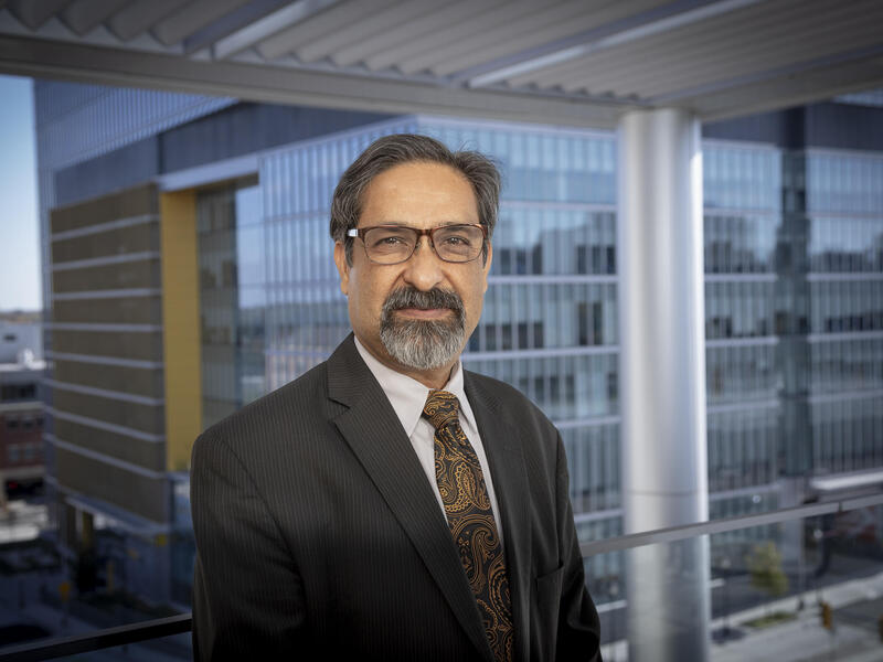 A photo of a man wearing a suit and tie from the chest up. He is standing in front of a window. 