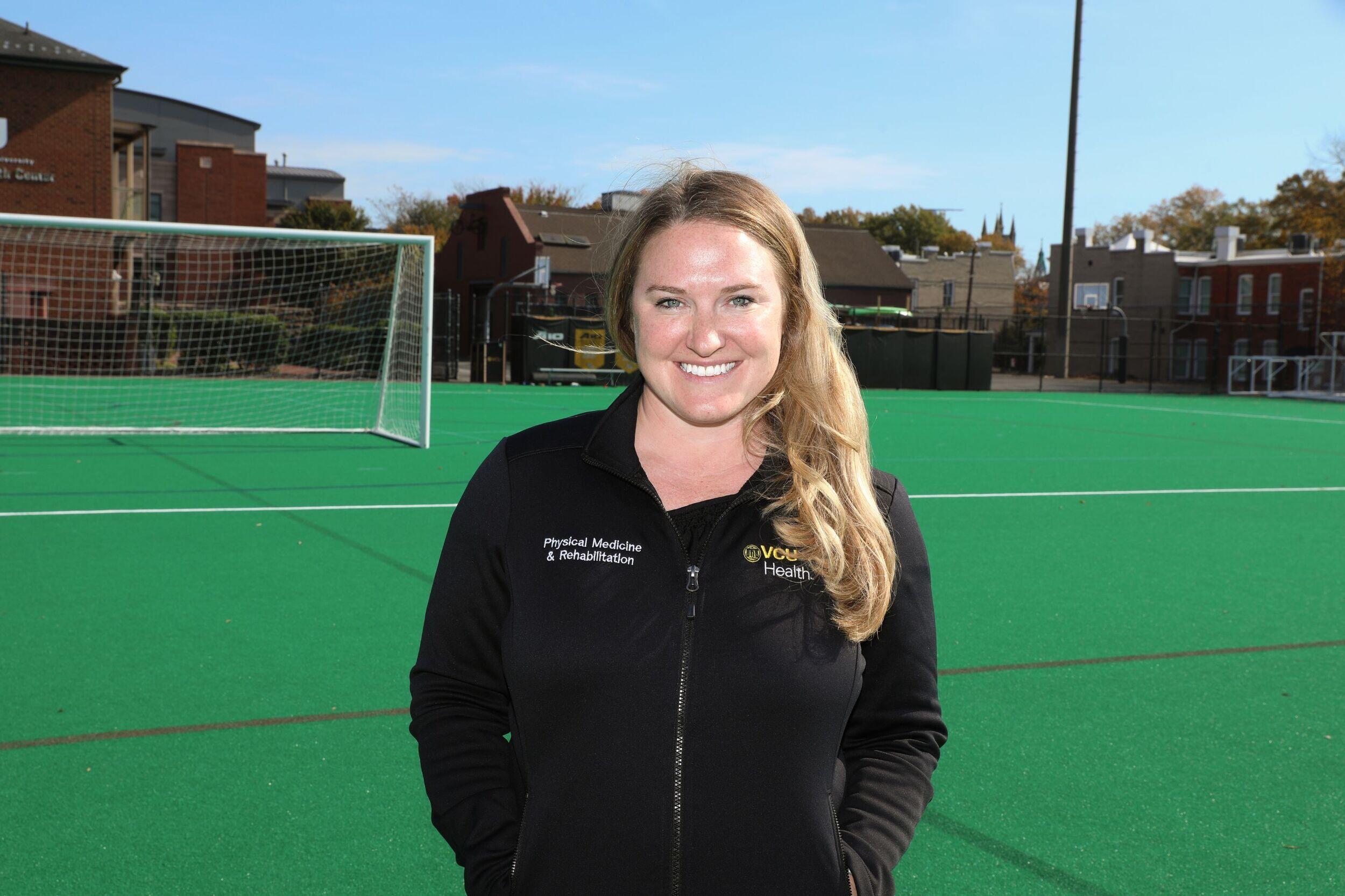 A photo of a woman from the waist up. She is standing on a soccer field. 