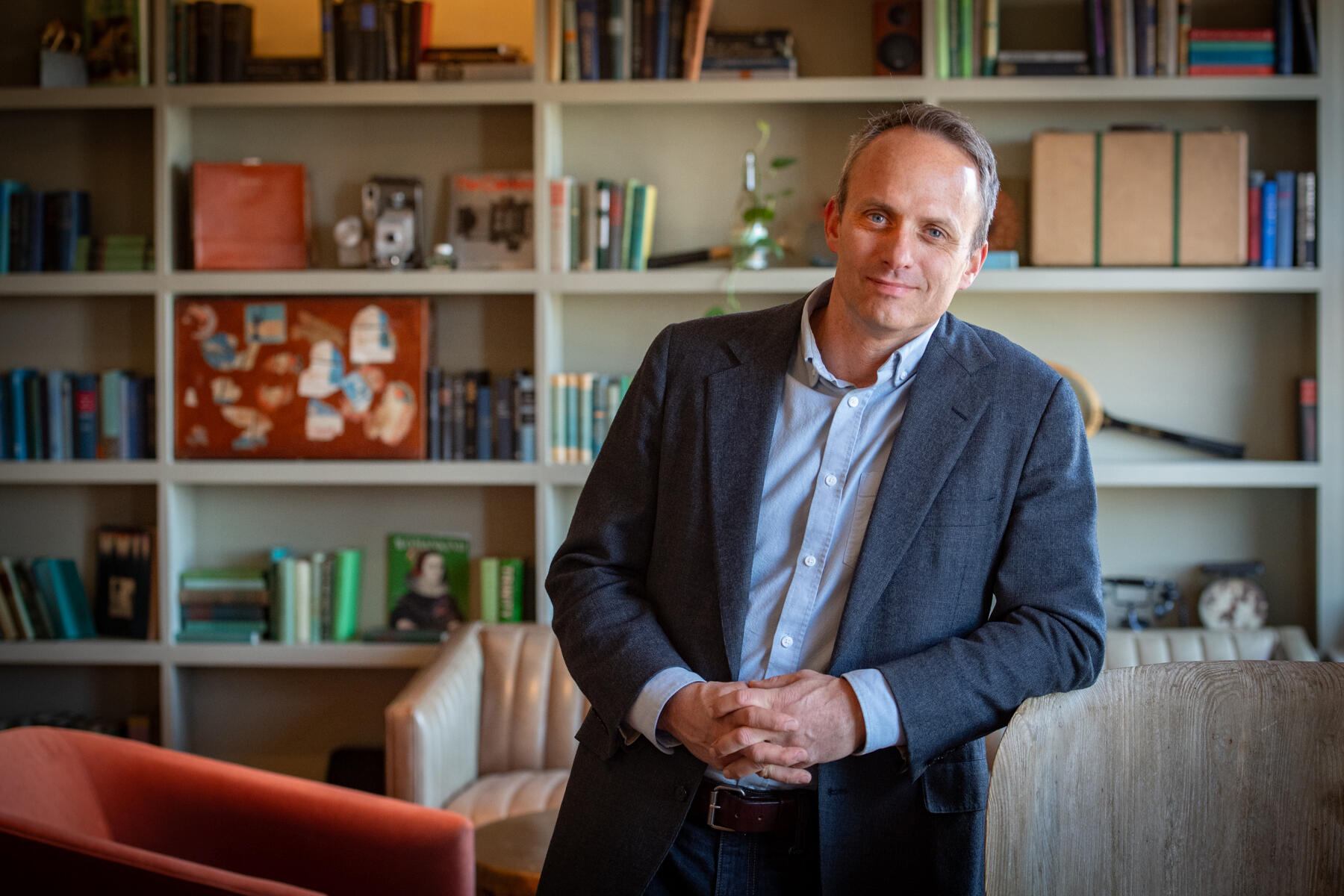 A photo of a man staning in front of a series of book shelves. He is leaning his right arm against a chair.