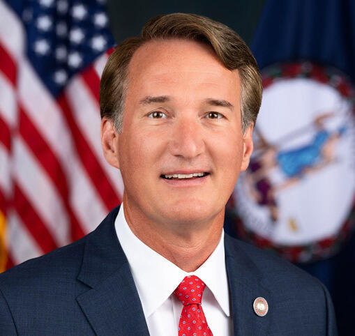A photo of a man wearing a suit and tie sitting in front of the U.S. and Virginia State flags. 