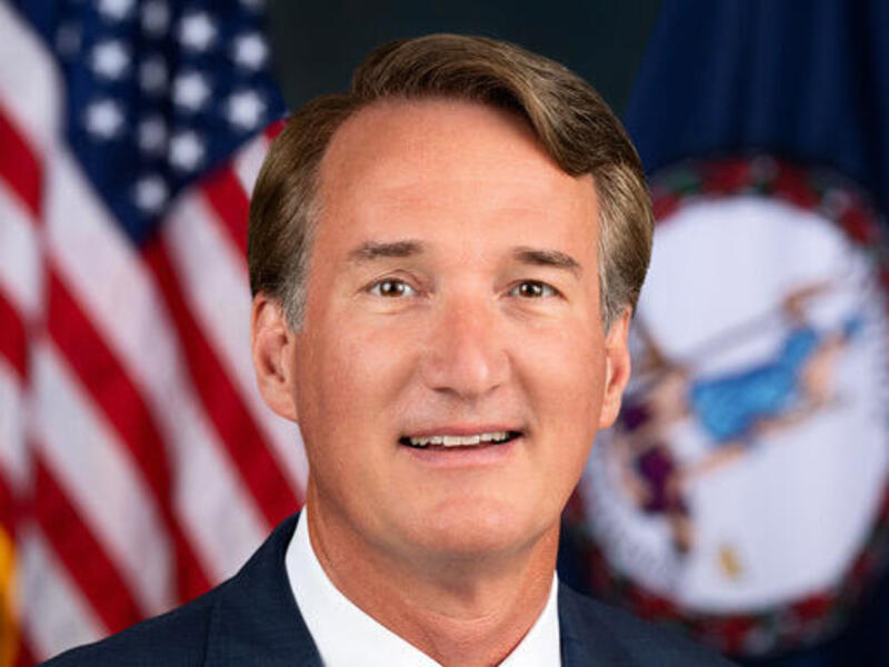 A photo of a man wearing a suit and tie sitting in front of the U.S. and Virginia State flags. 