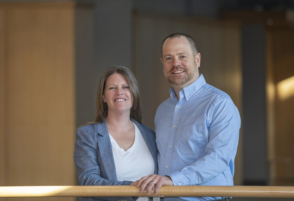 A woman standing to the left of a man. Her left hand is touching his right hand, and both hands are resting on a railing. 