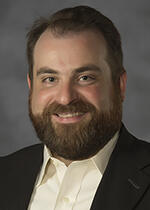 A portrait of a man with a beard wearing a white button up shirt and a black suit jacket. 