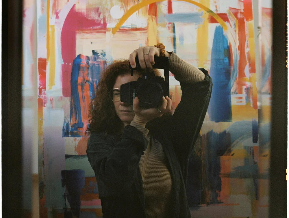 A photo of a woman taking a picture of herself in a mirror. Behind her is a multi-colored abstract painting. 