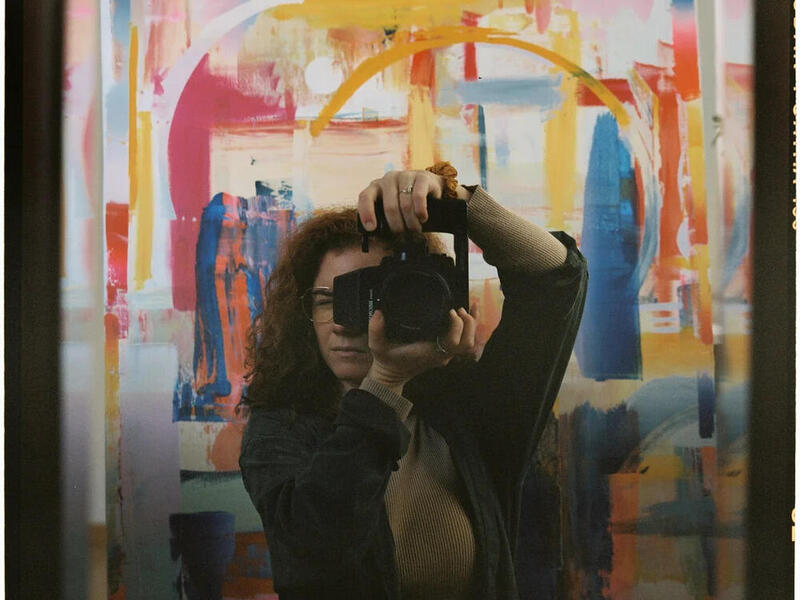 A photo of a woman taking a picture of herself in a mirror. Behind her is a multi-colored abstract painting. 