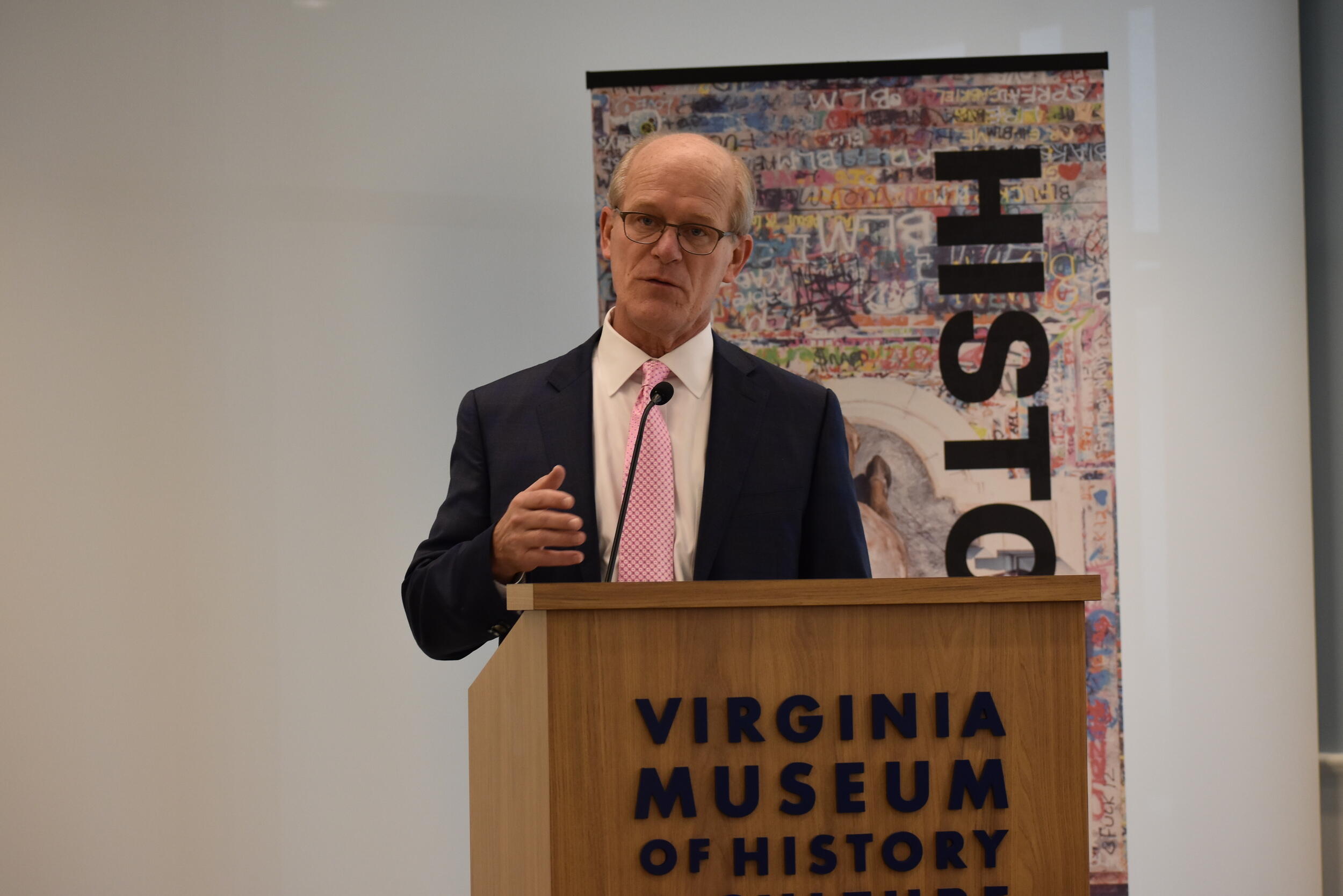 A photo of a man speaking behind a podium. The front of the podium says \"VIRGINIA MUSEUM OF HISTORY\" in black letters. 