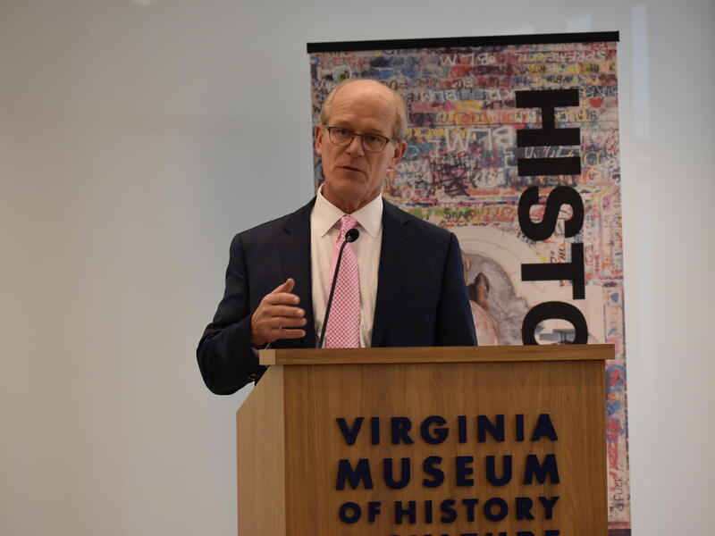 A photo of a man speaking behind a podium. The front of the podium says \"VIRGINIA MUSEUM OF HISTORY\" in black letters. 