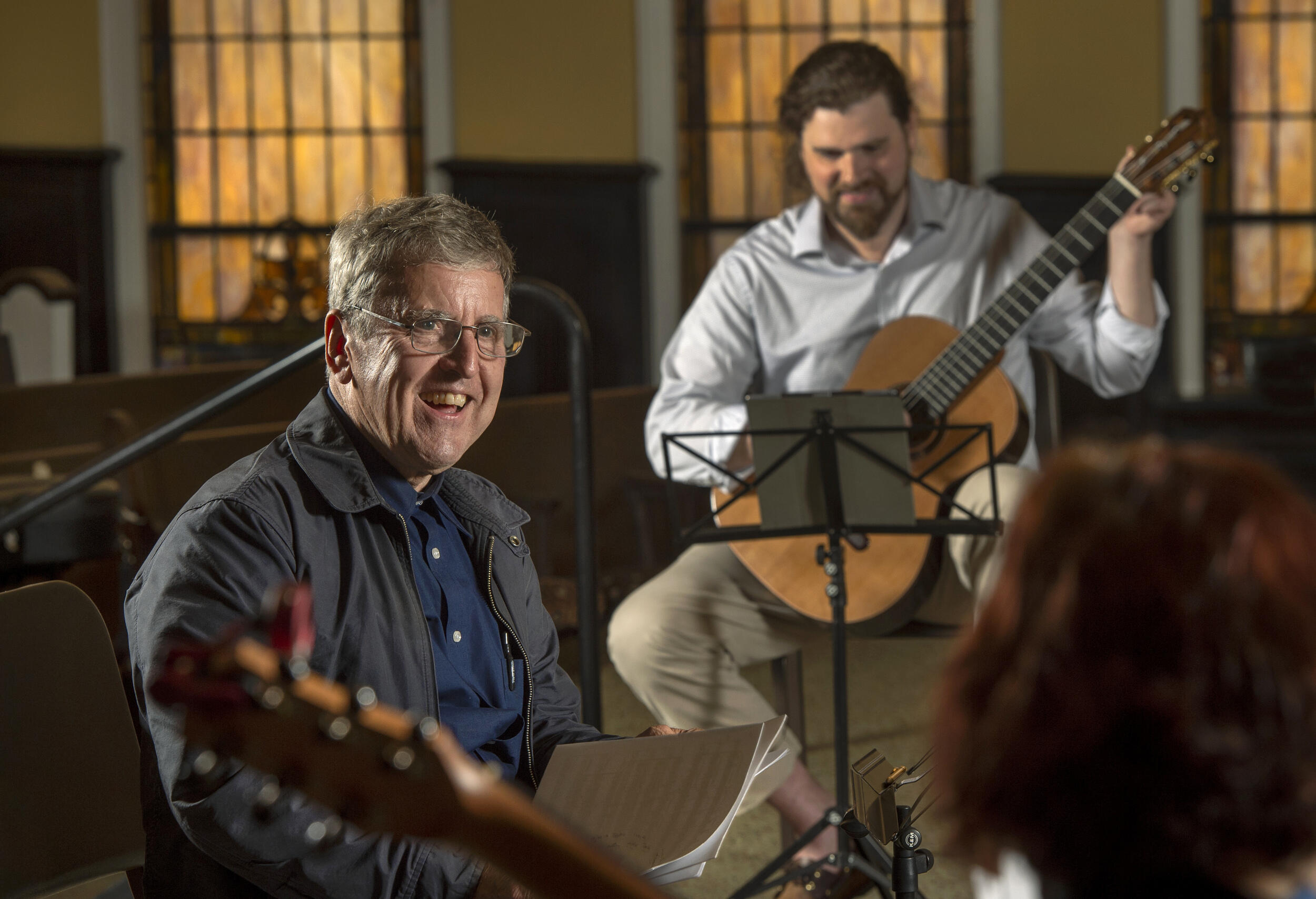 A photo of a man sitting on a chair holding sheet music. Behind him to the right is another ma sitting down holding a guitar. 