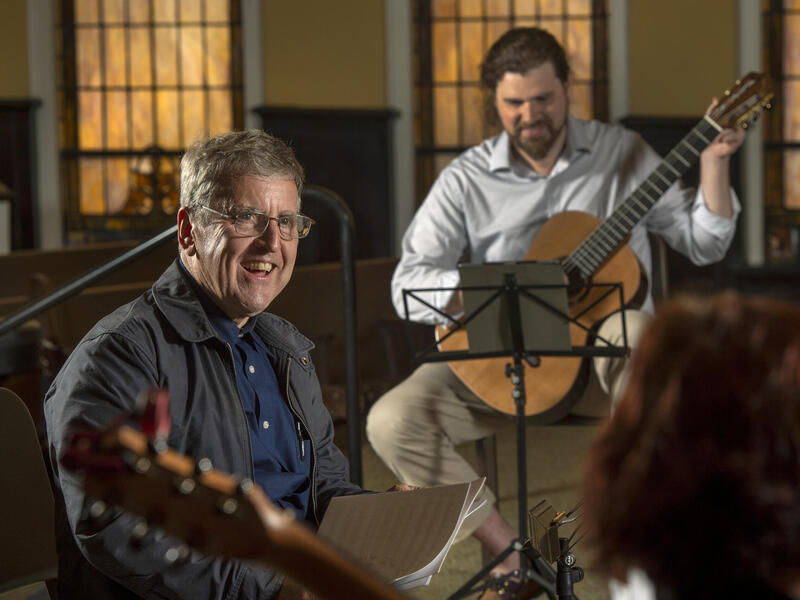 A photo of a man sitting on a chair holding sheet music. Behind him to the right is another ma sitting down holding a guitar. 