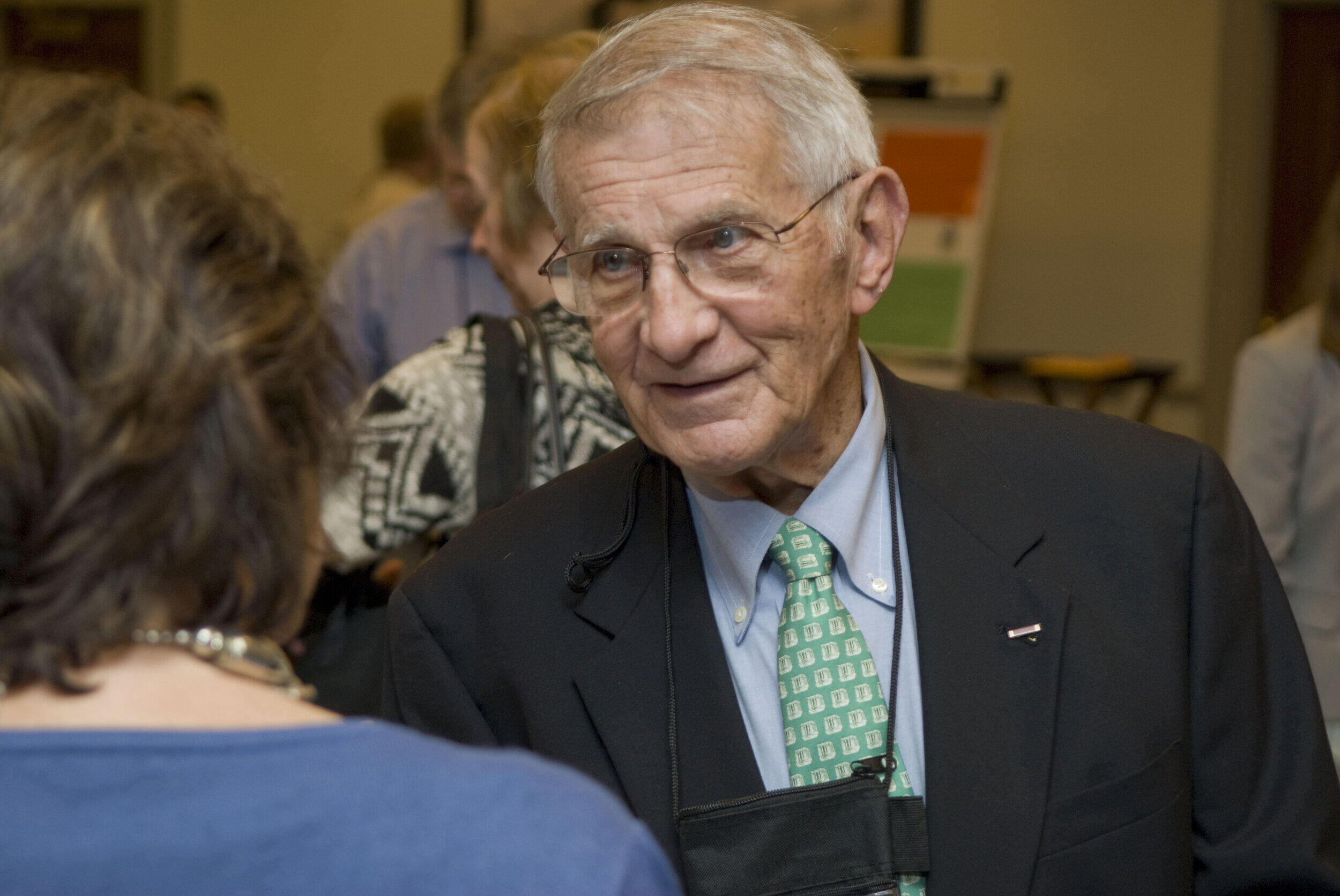 An old man wearing glasses, a black sports jacket, a greet tie and a light blue shirt. 