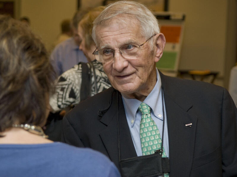 An old man wearing glasses, a black sports jacket, a greet tie and a light blue shirt. 
