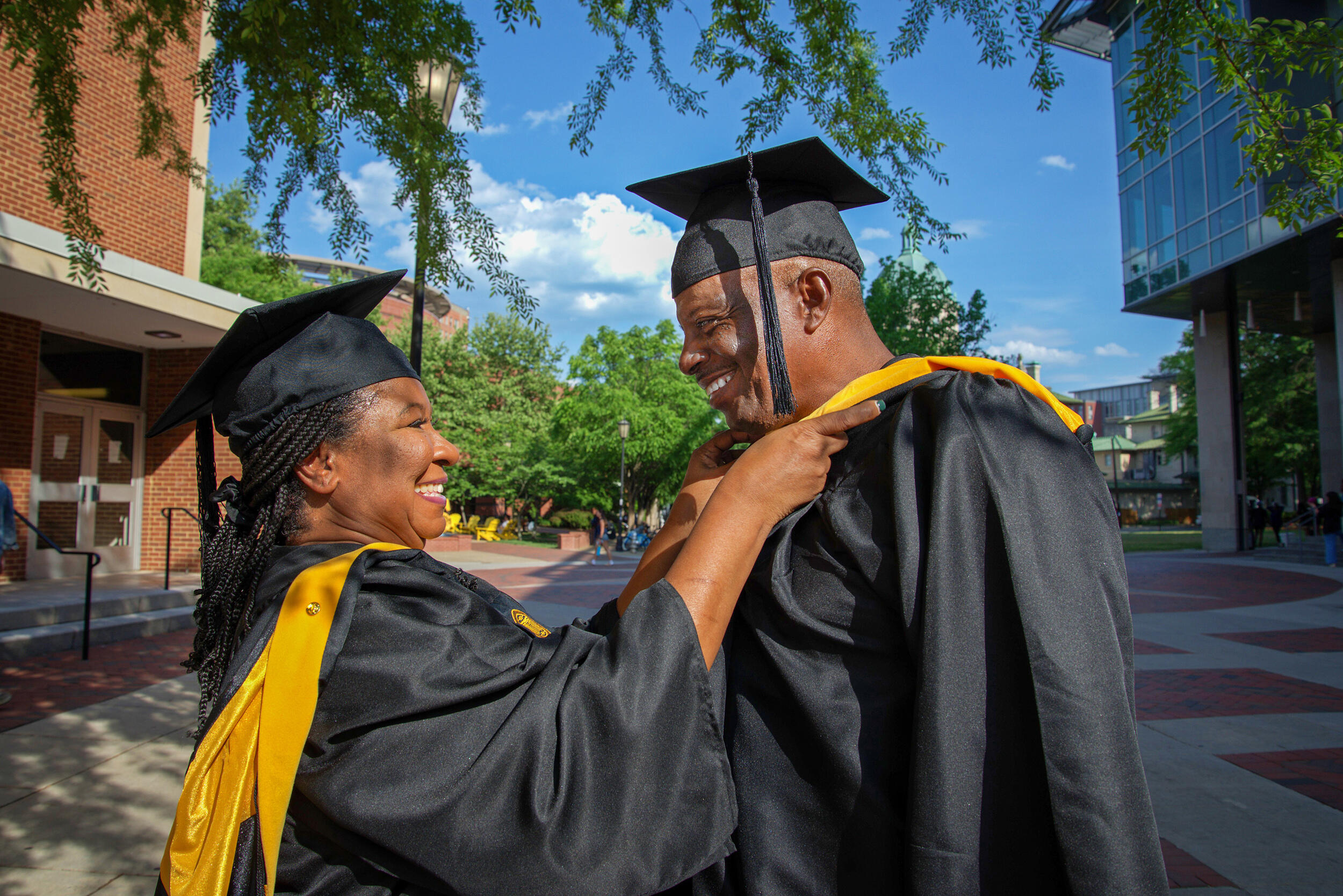 A photo of a man and woman wearing graduation caps and gowns. The woman is placing a stole arond the man's neck. 