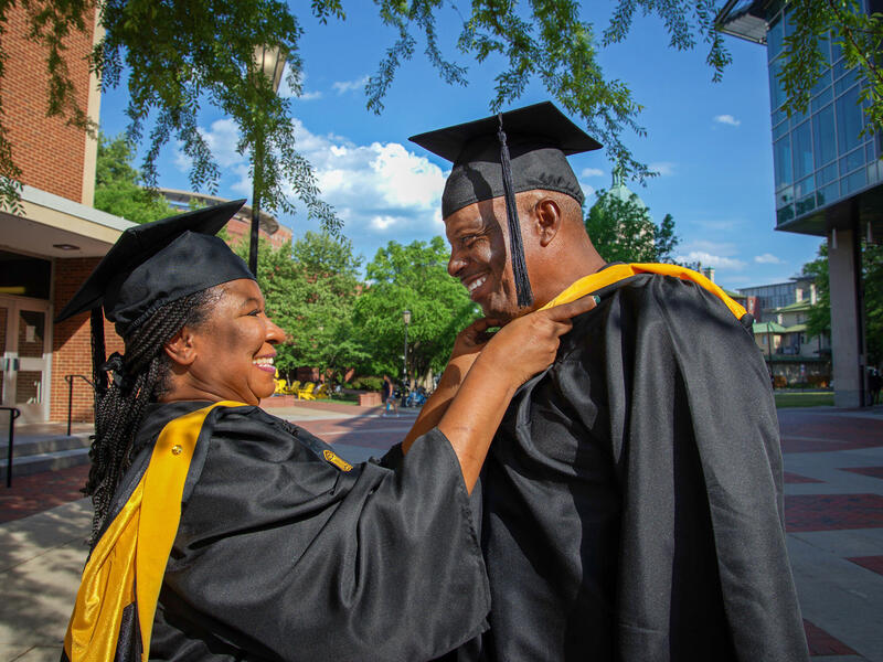 A photo of a man and woman wearing graduation caps and gowns. The woman is placing a stole arond the man's neck. 
