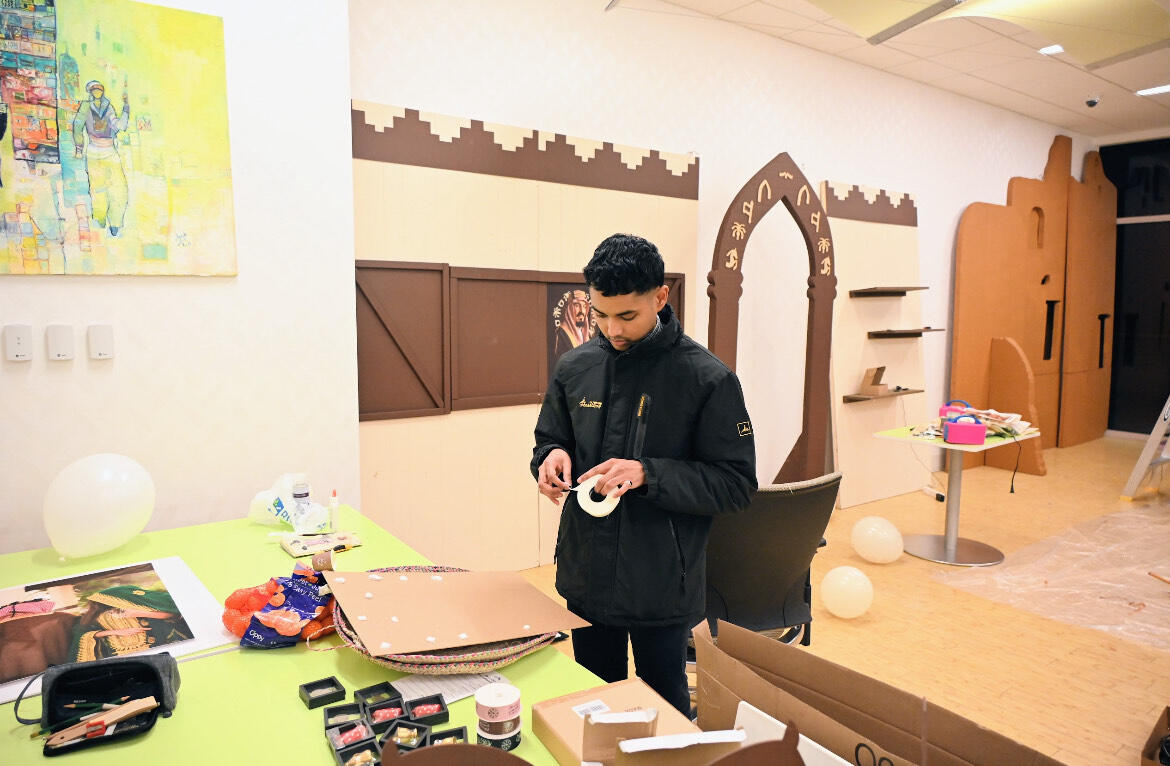 A photo of a man holding a roll of tape standing in front of a table covered in objects. 