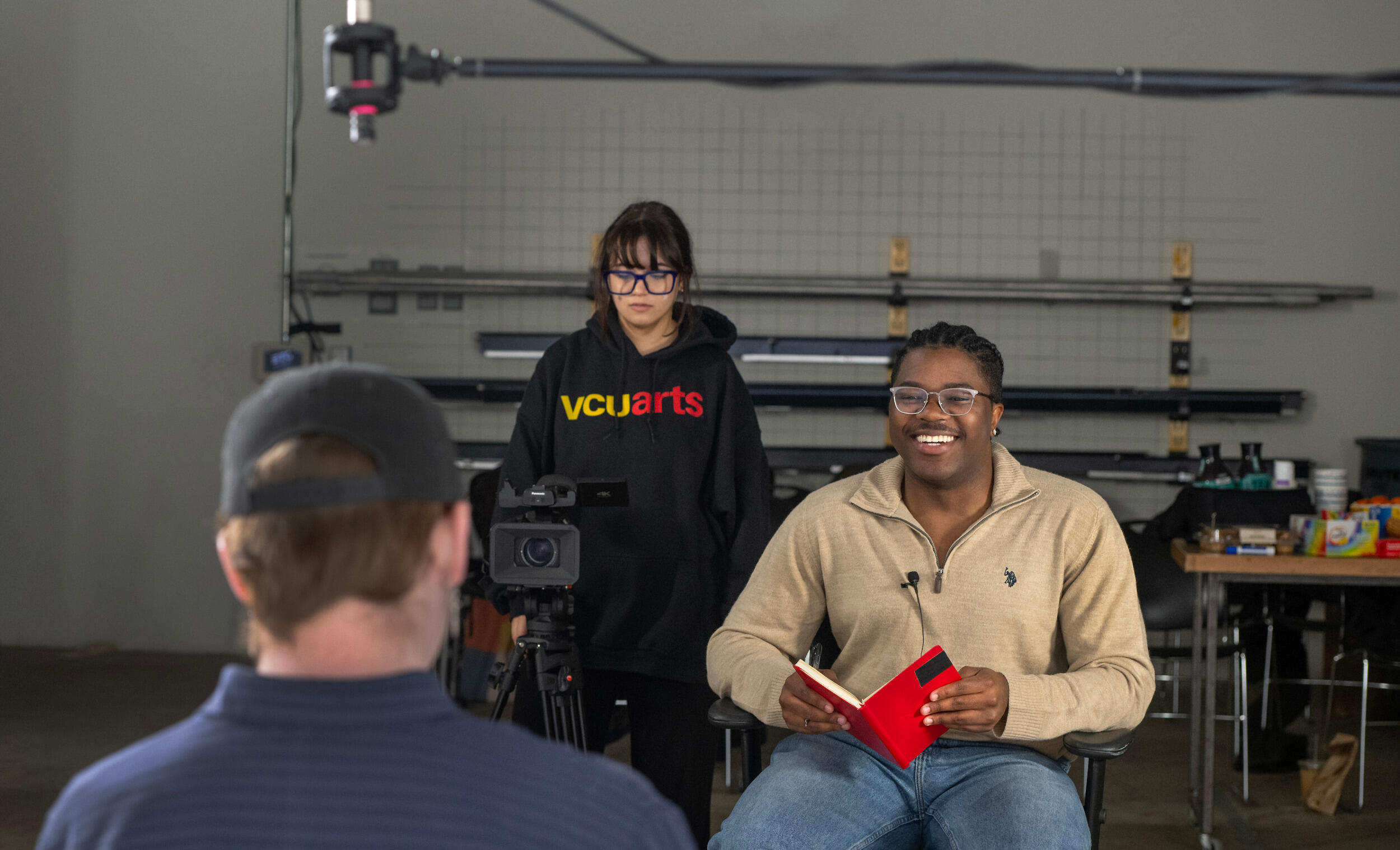 A man facing the camera smiles with a book open in front of him. A woman stands slighlty behind him with a video camera. They are facing a man in the foreground who has his back to the camera.