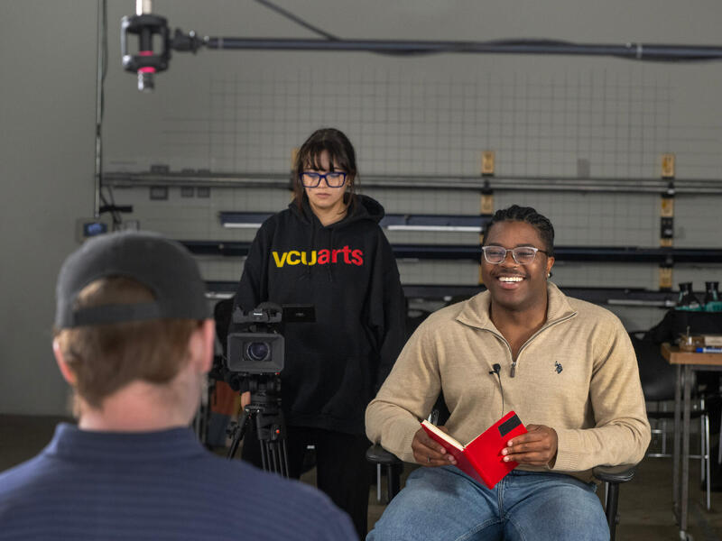 A man facing the camera smiles with a book open in front of him. A woman stands slighlty behind him with a video camera. They are facing a man in the foreground who has his back to the camera.