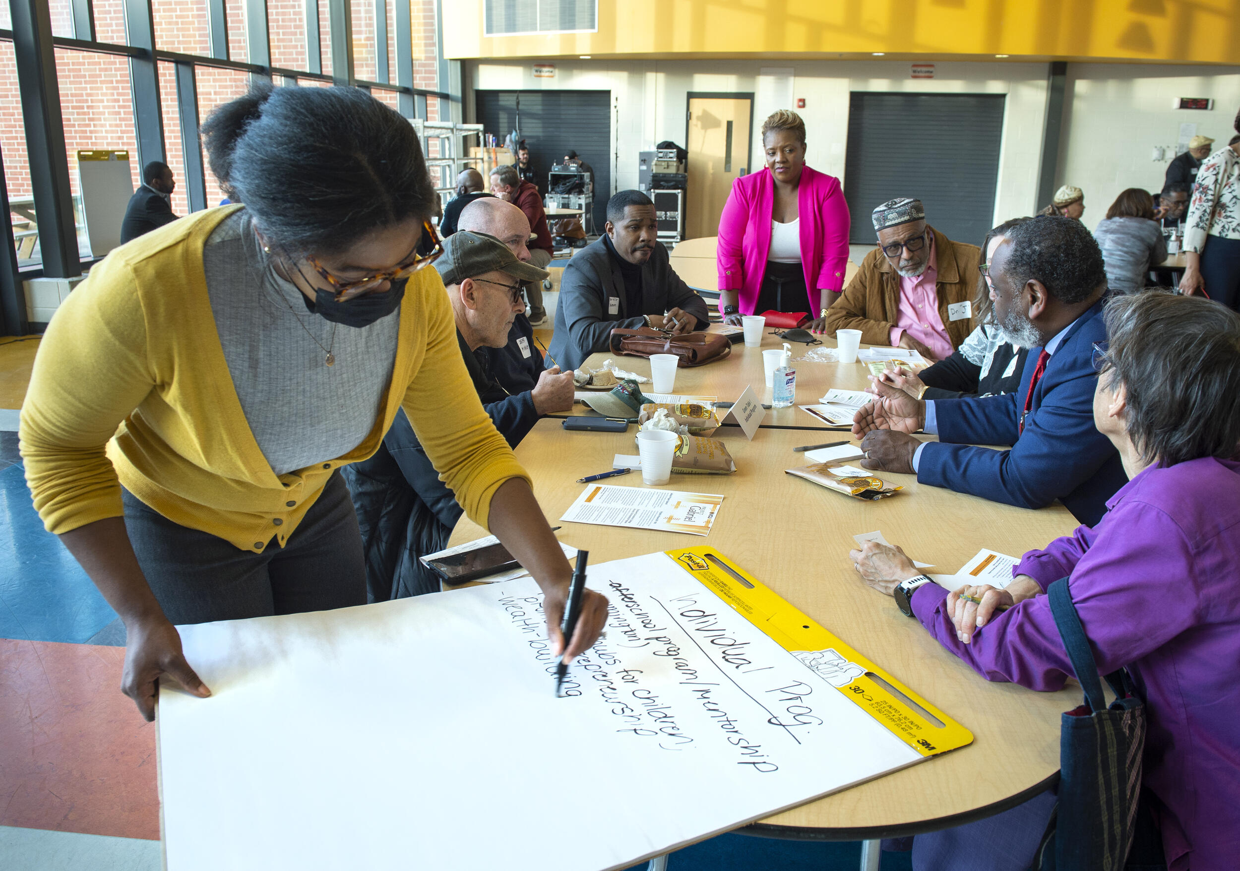 Six people are sitting at a table and two women are standing at the ends of it. At the end of the table closest to the front, a woman is writing on a large pad of paper. 