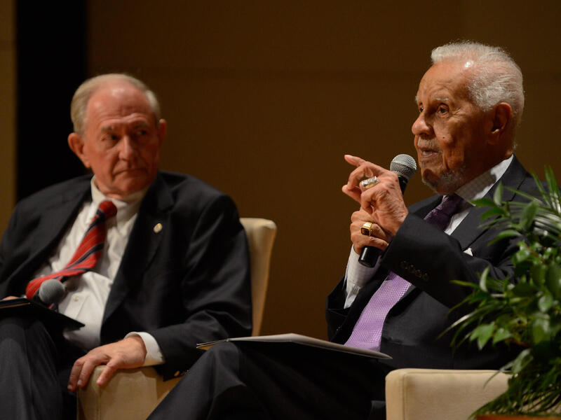 A photo of two men sitting in arm chairs. The man on the right is speaking into a microphone. 