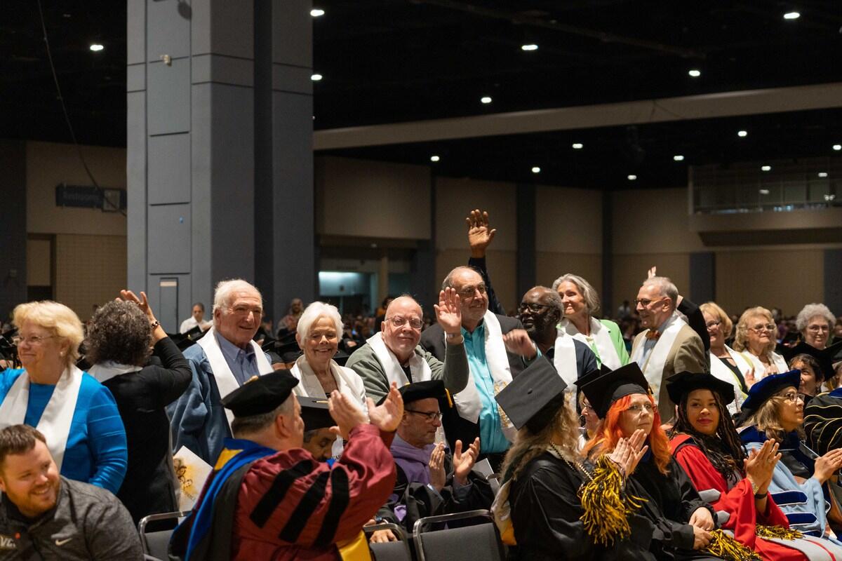 A photo of 12 older adults standing and waving while people in graduation caps sitting in front of the group clap. 