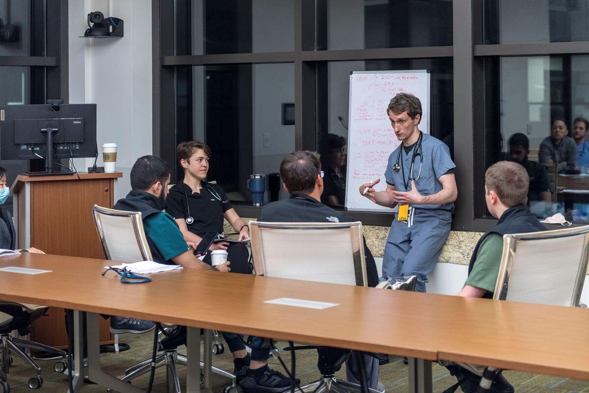 A photo of a man talkng to four other people sitting in a semi-circle of chairs. 