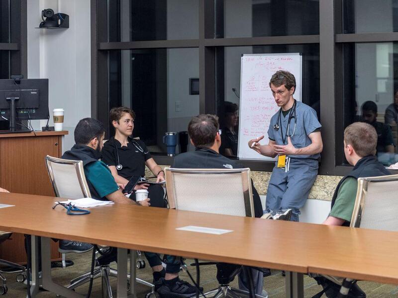 A photo of a man talkng to four other people sitting in a semi-circle of chairs. 