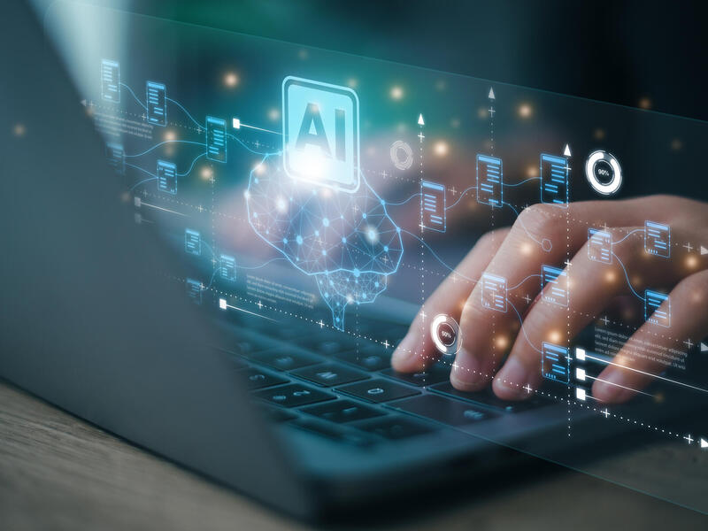 A photo of a hand typing on a laptop. In front of the laptop screen is a projection that shows an illustration of a brain and text that reads \"AI\"