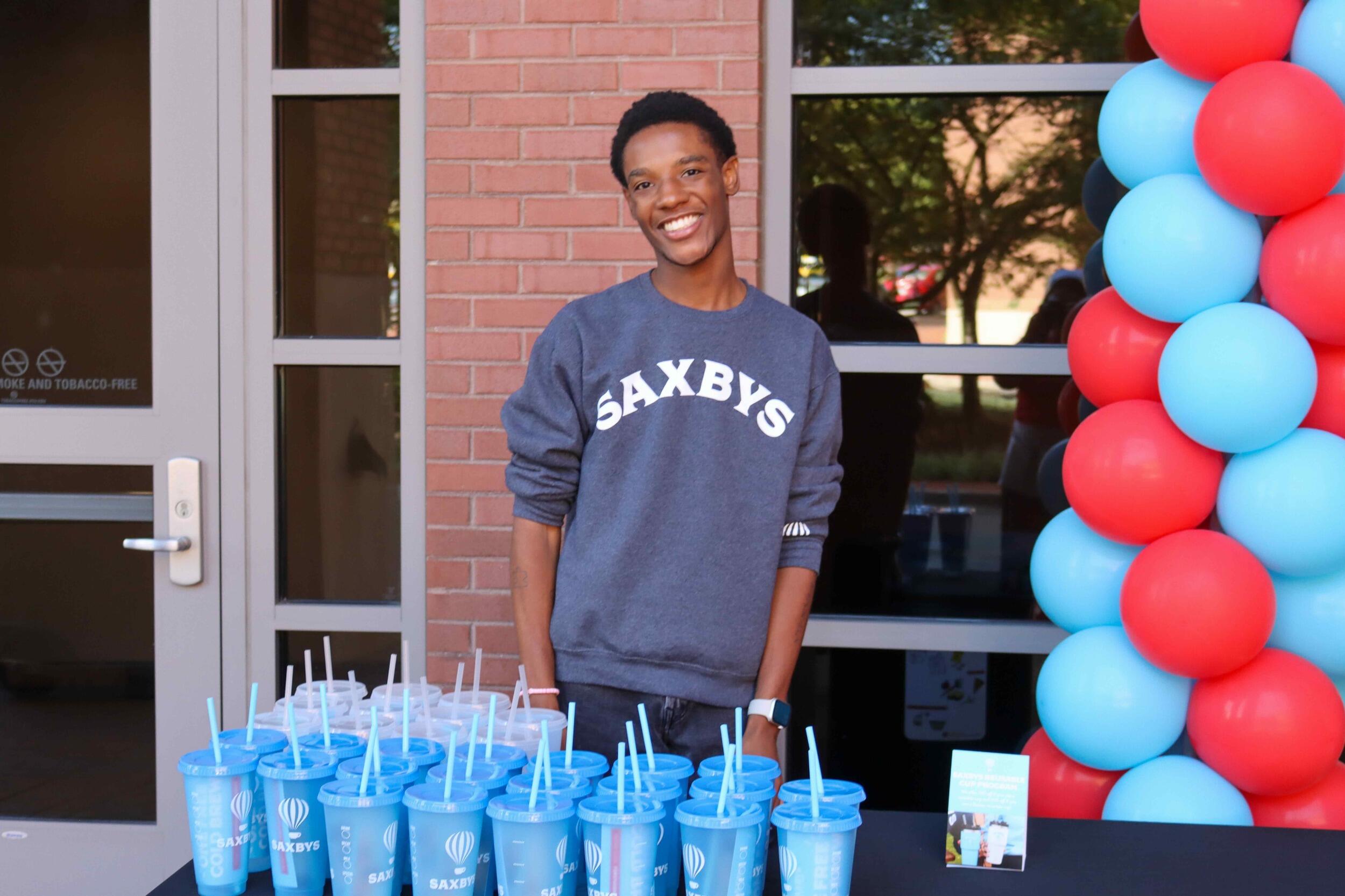A photo of a man standing behind a table that has light blue tumblers that says \"Saxbys\" on them in white text 