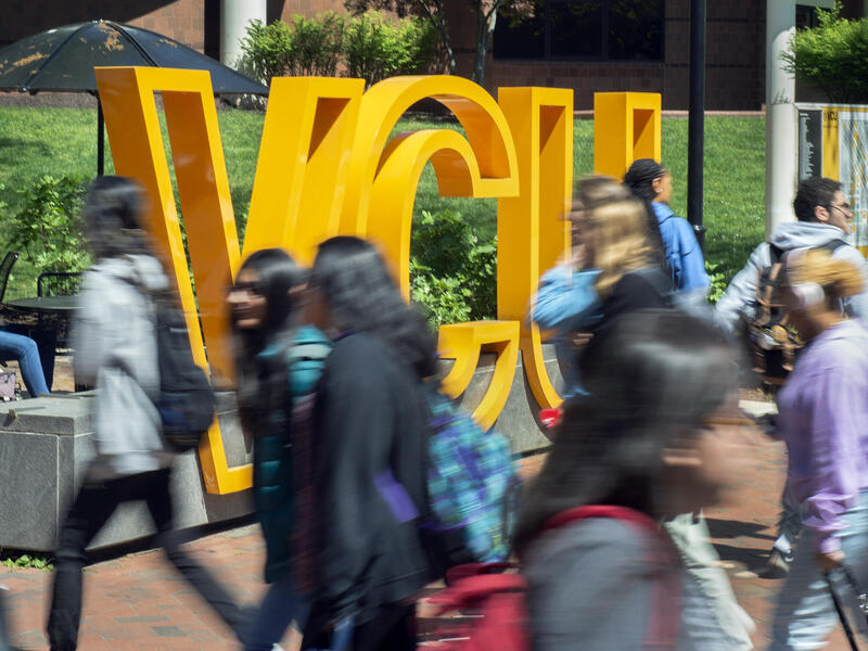 A photo of people walking past a sign that spells out \"VCU\" in large yellow letters