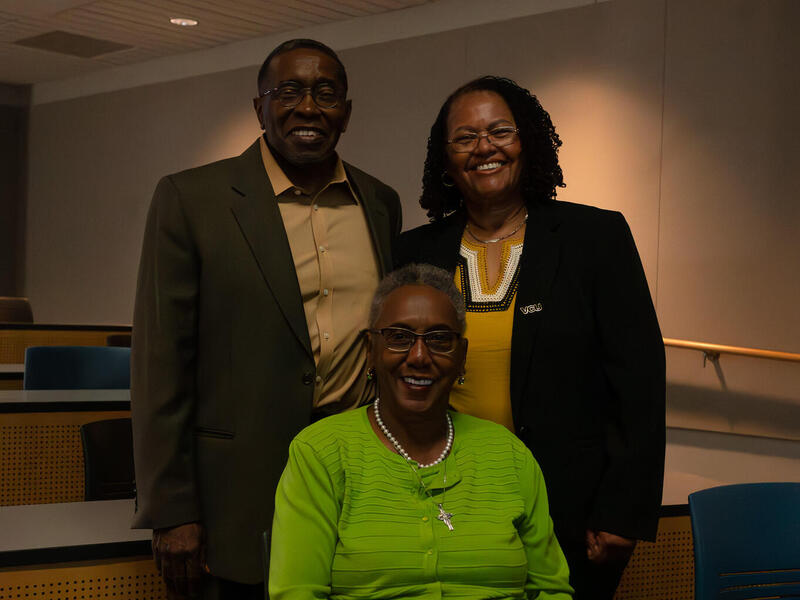 One woman sits at a table in a green jacket and a man and a woman stand behind her.