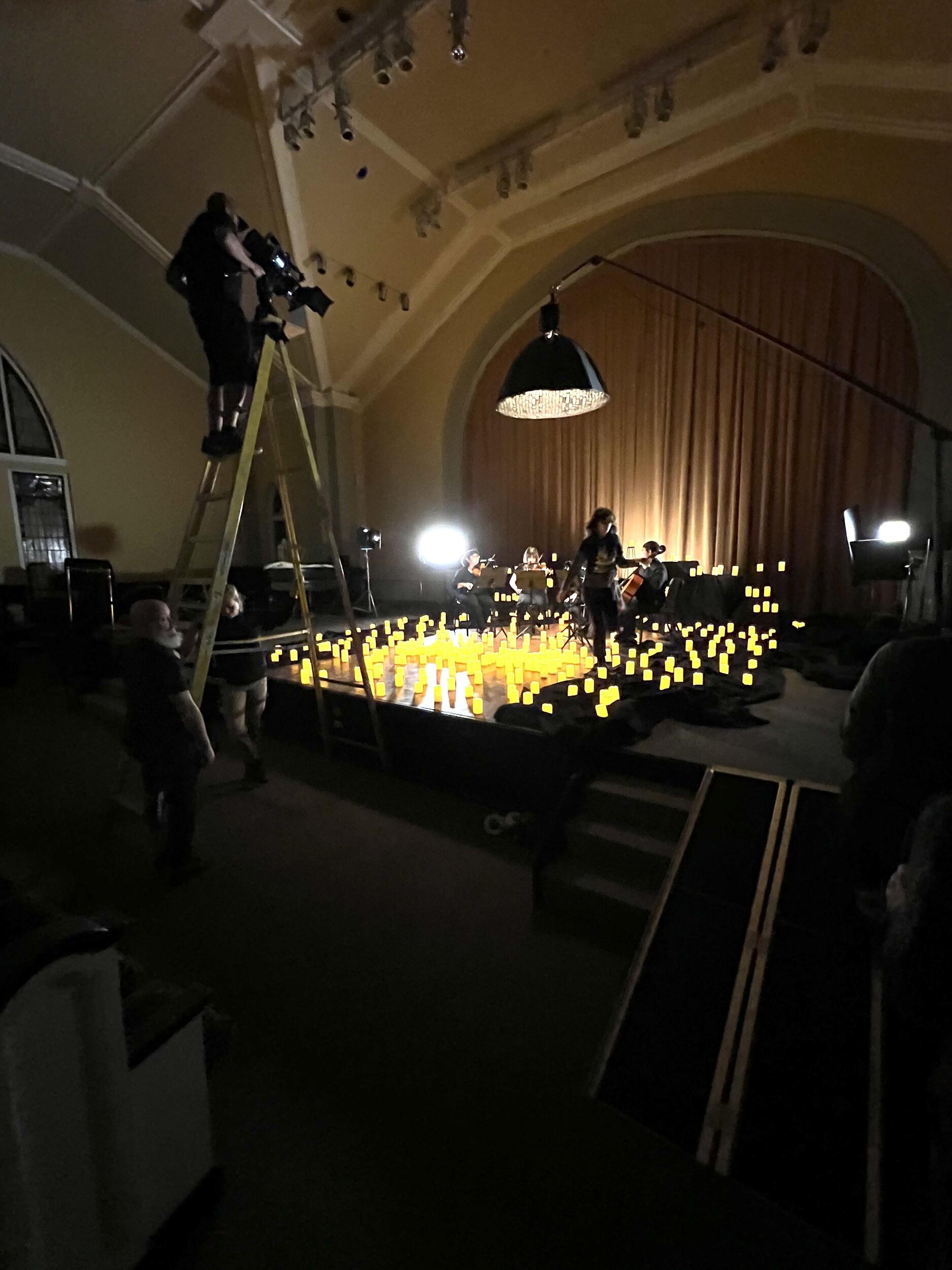 A cameraman standing on a ladder filming a stage lit with hundreds of candles as a string quartet plays.
