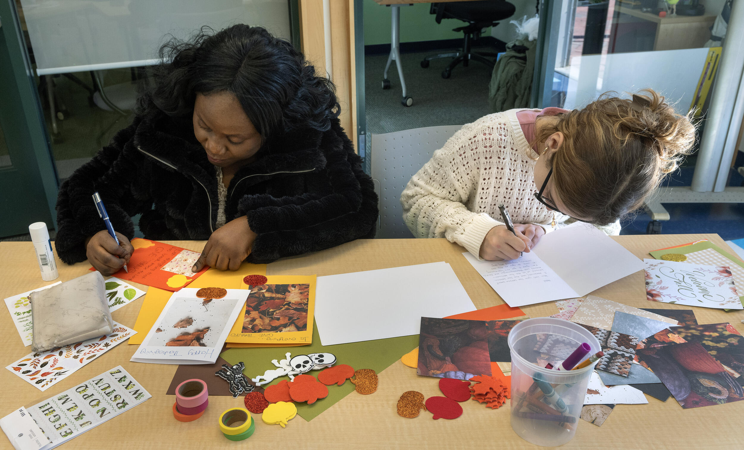 A photo of two women sitting at a table covered in stamps, stickers, and paper. The two women are writing inside of cards. 