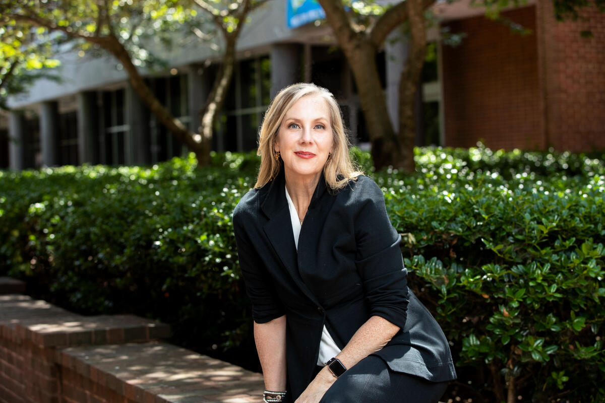 A photo of a woman sitting outside in front of bushes and trees that are next to a building. 