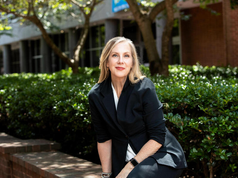 A photo of a woman sitting outside in front of bushes and trees that are next to a building. 