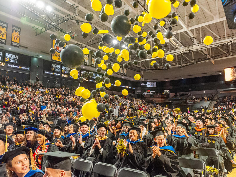 A photo of black and yellow balloons falling down on a group of students wearing graduation cap and gowns. The students are sitting in rows of chairs. Around the students are bleachers filled with people sitting in them.