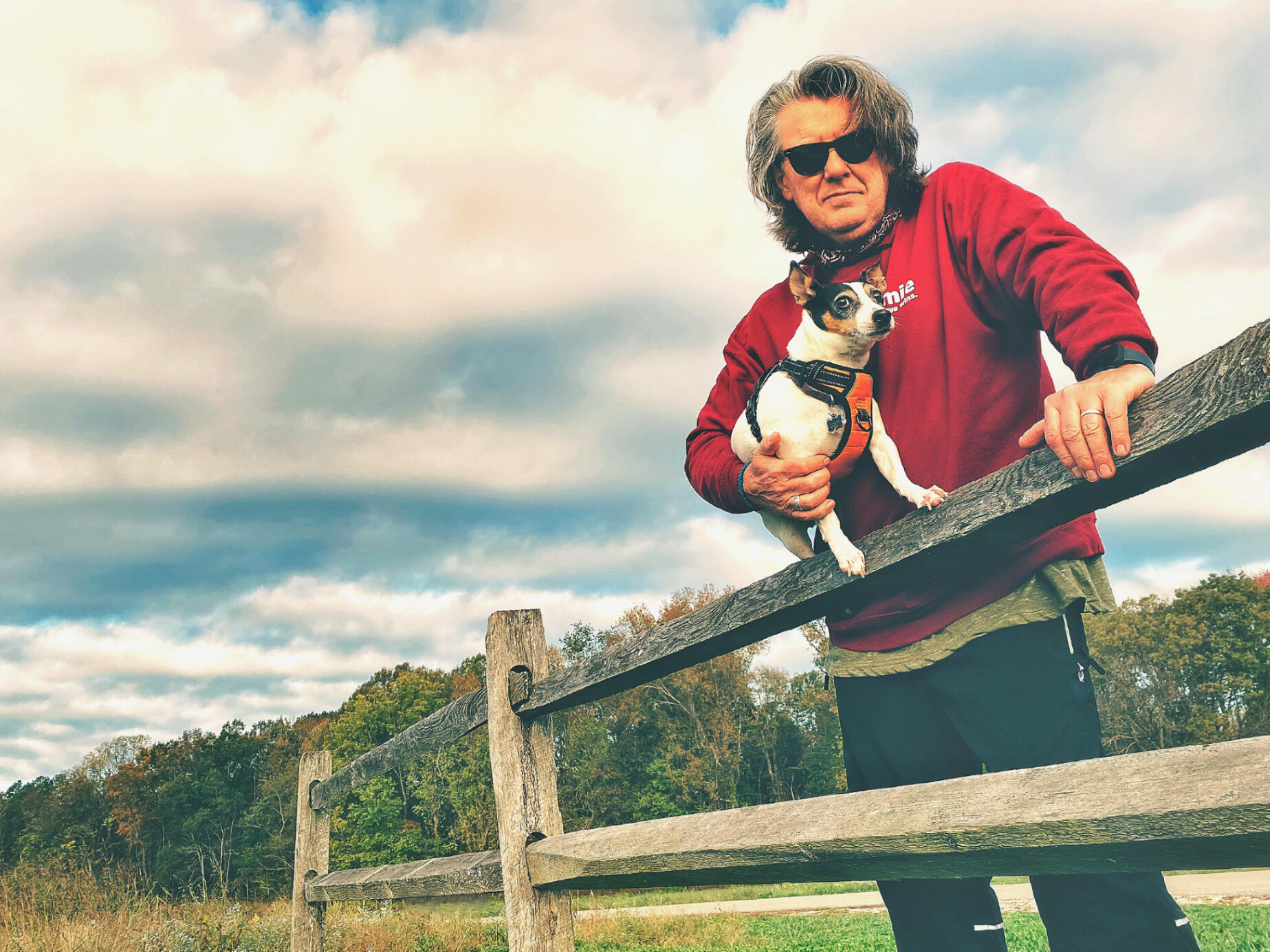 A man leans on a fence while holding a small dog with one arm.