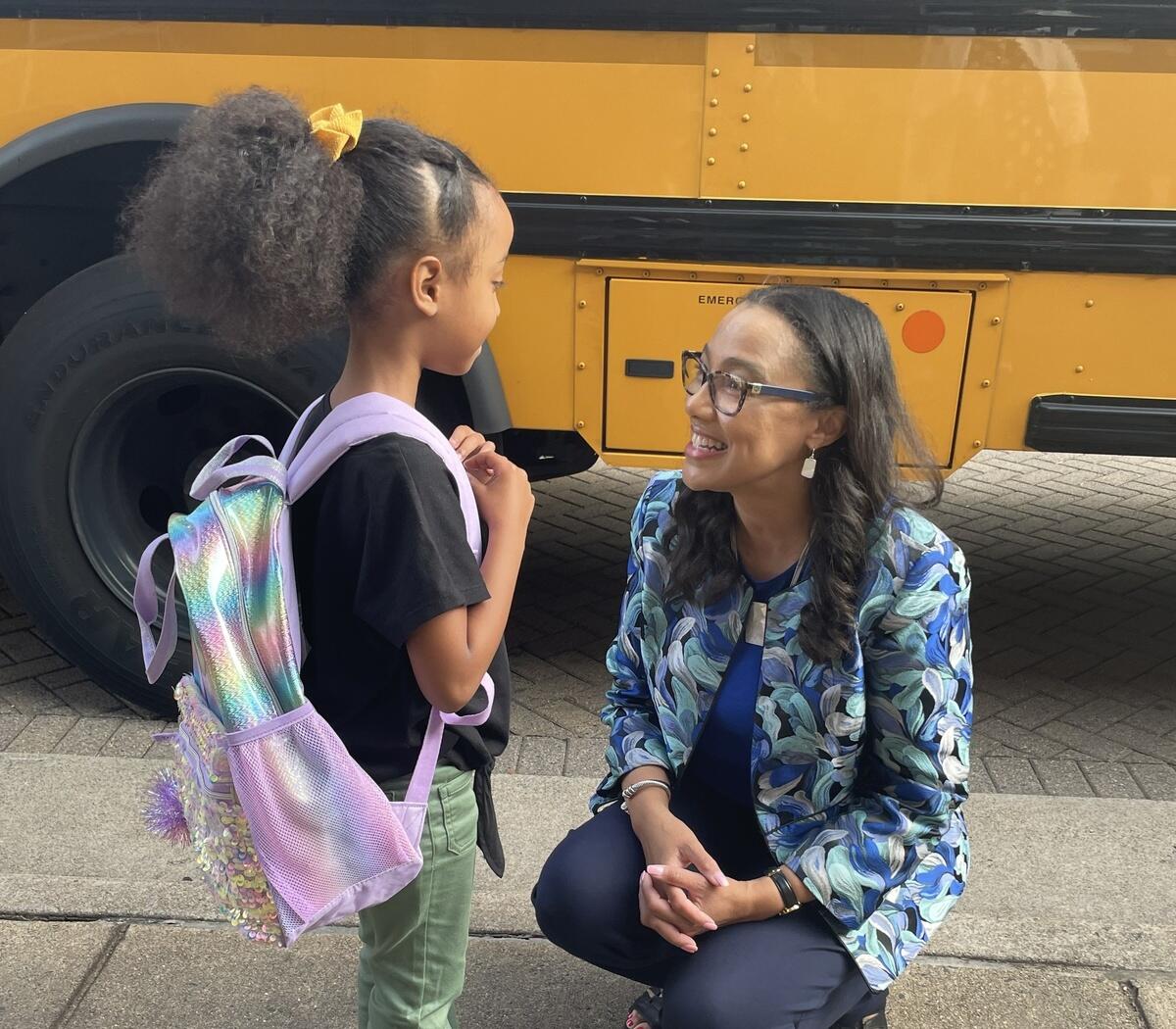 A photo of a woman kneeling next to a young girl wearing a purple backpack. They are both in front of a yellow school bus. 