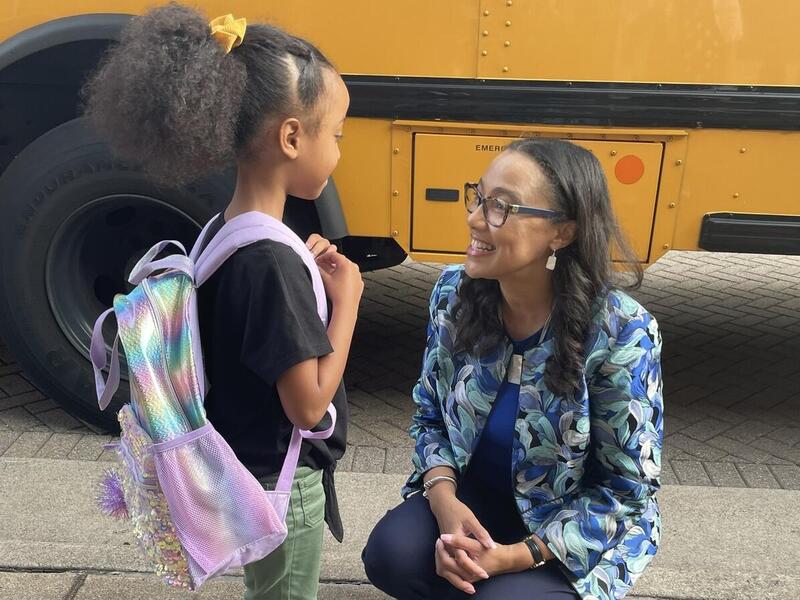 A photo of a woman kneeling next to a young girl wearing a purple backpack. They are both in front of a yellow school bus. 
