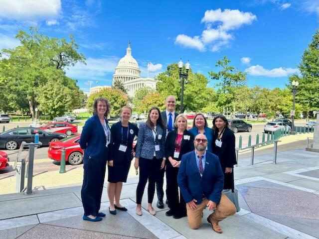 A photo of seven people standing and one man kneeling on a sidewalk. Behind the group is the U.S. Capitol. 