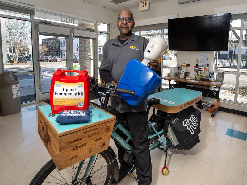 A photo of a man standing over a bike. He is holding a CPR dummy. On the front of the bike is a wooden box with a blue top. Sitting on top of the box is a emergency opioid kit.