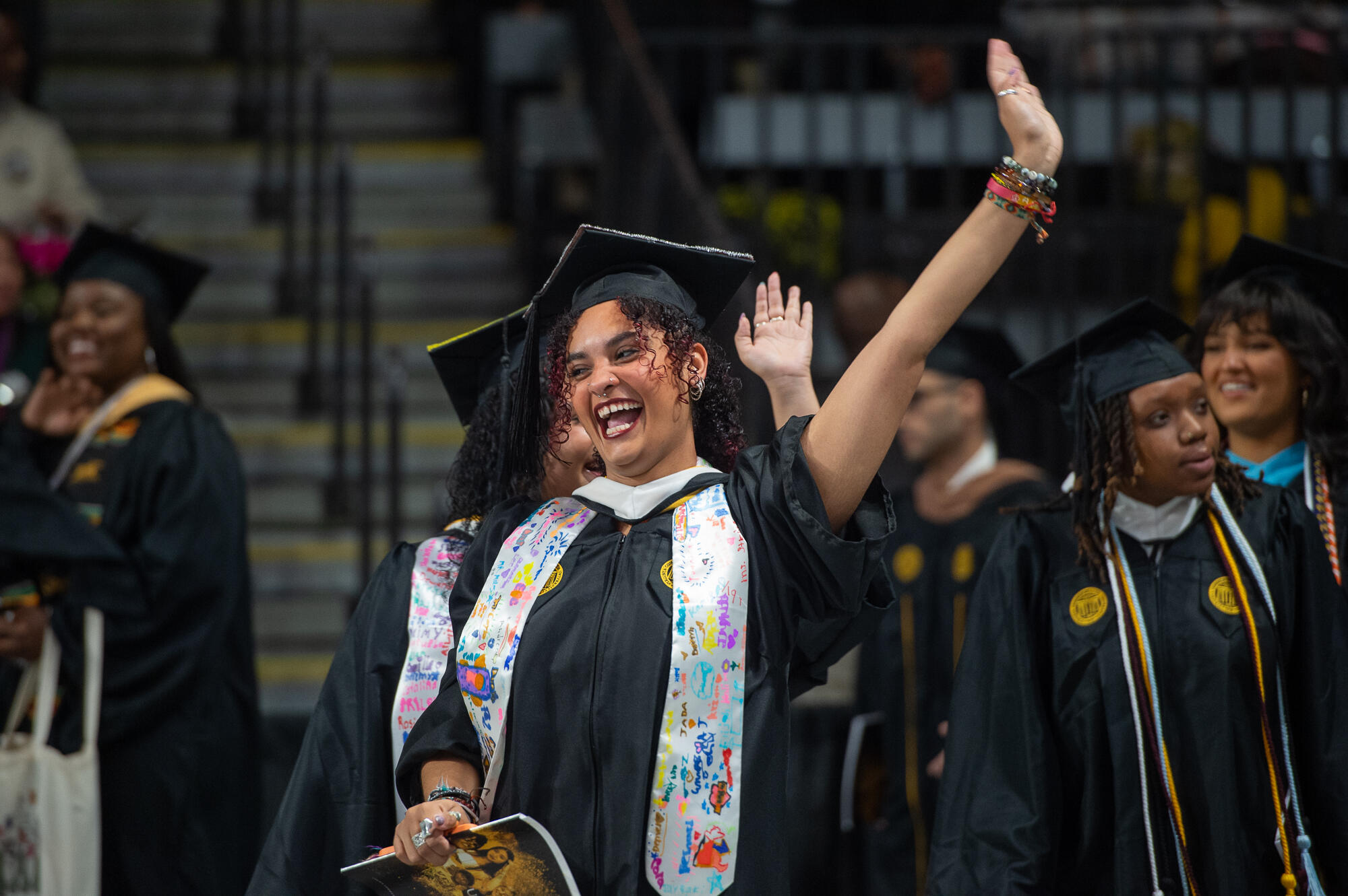 A graduate in cap and gown raises her hand and smiles broadly.