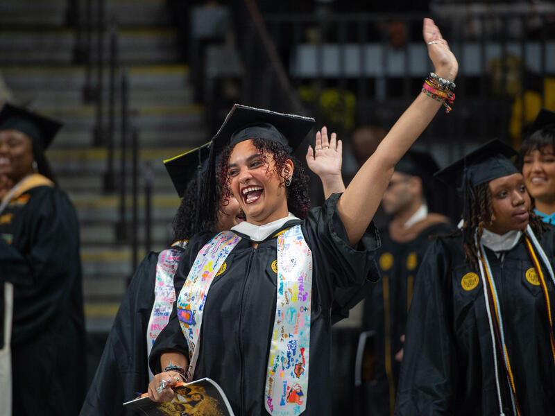 A graduate in cap and gown raises her hand and smiles broadly.