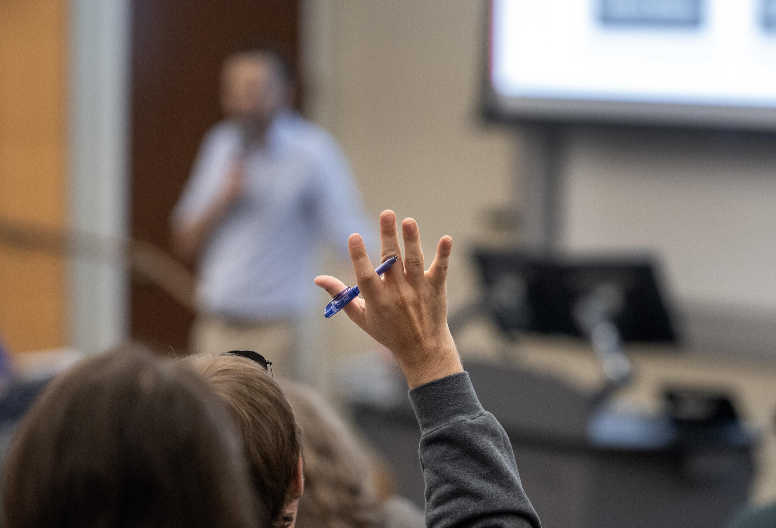 A photo of a person raising their hand while the hand also holds a pen. In the background is a blurred image of a person holding a microphone. 