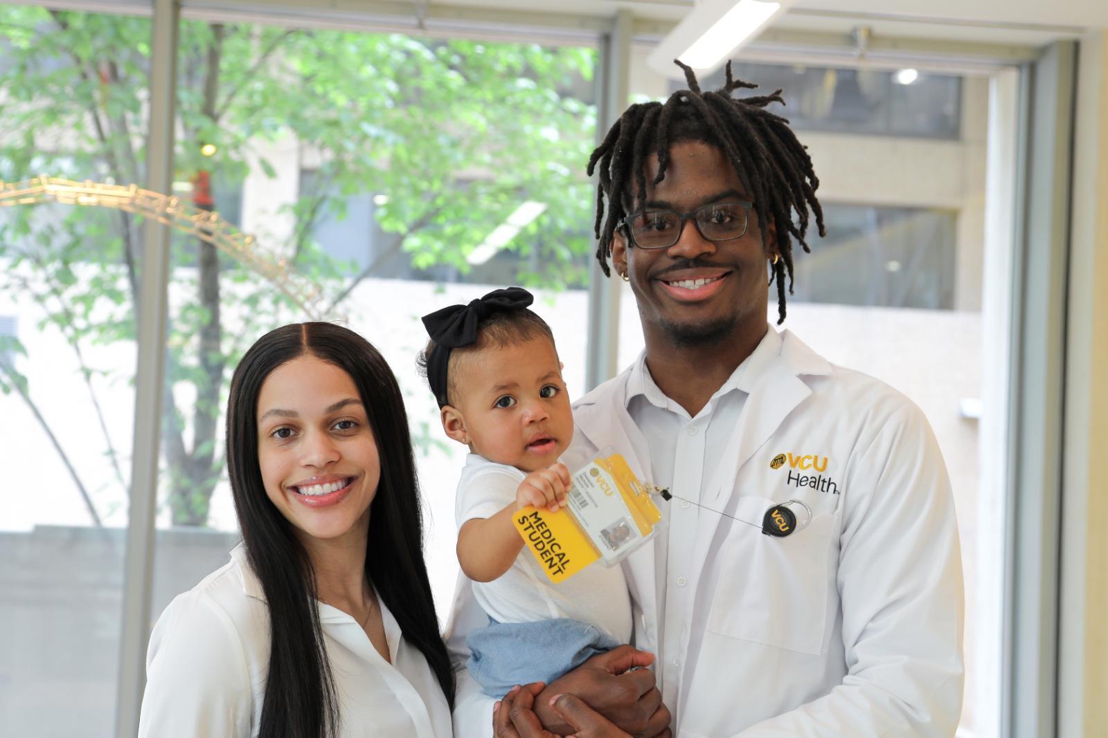 A photo of a man and woman standing next to each other. The man is holding a baby, who is playing with his ID badge