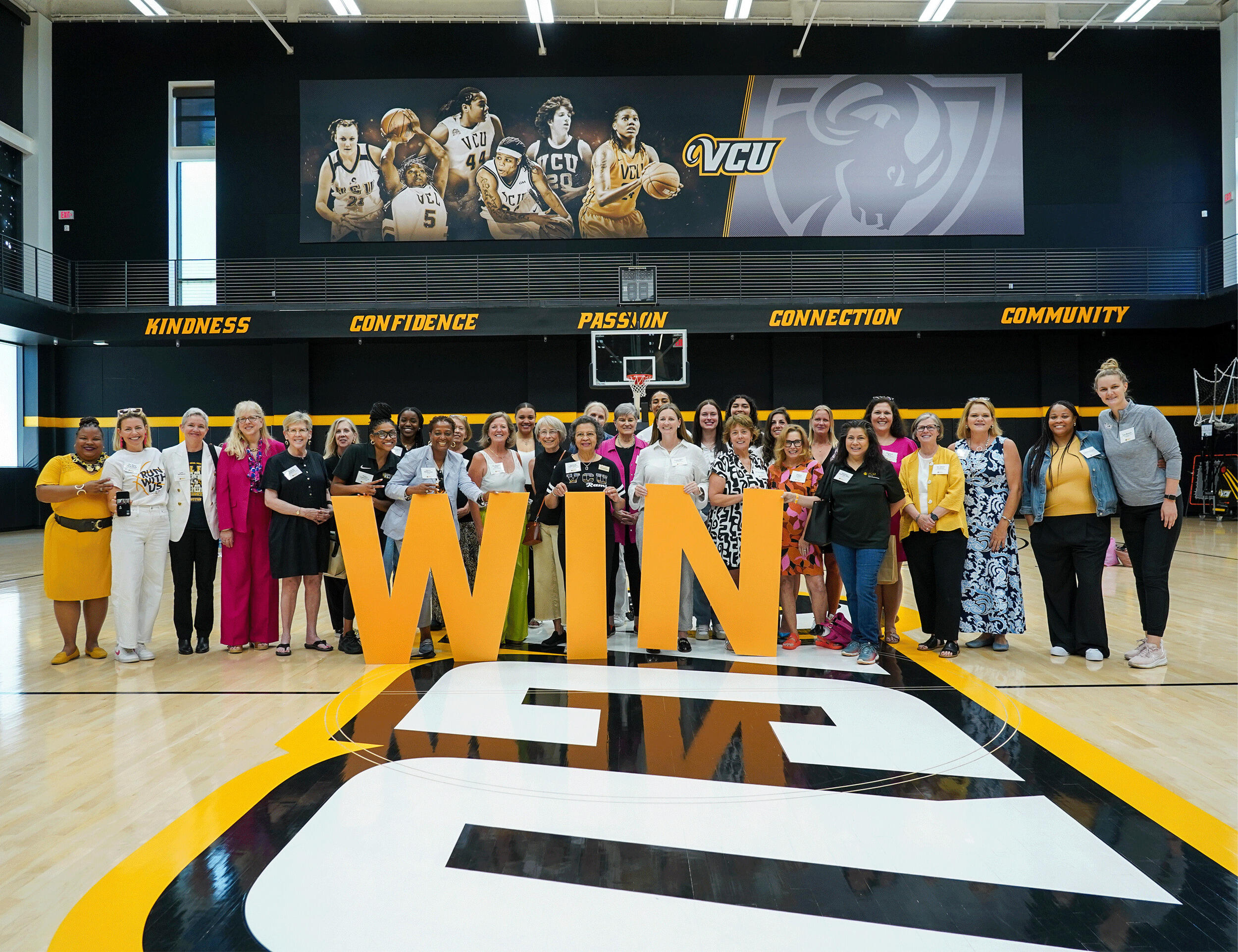 A photo of 30 women standing on a basketball court. The women are standing behind giant yellow letters that spell out \"WIN.\"