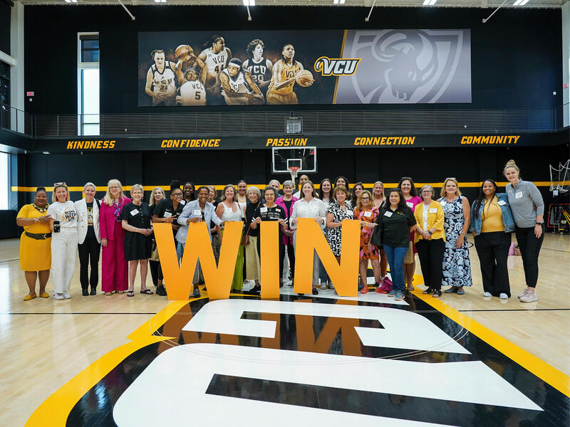 A photo of 30 women standing on a basketball court. The women are standing behind giant yellow letters that spell out \"WIN.\"