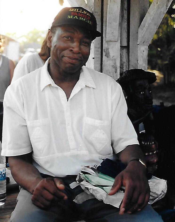 A man sitting and smiling at the camera. He is wearing a hat and holding assorted papers.